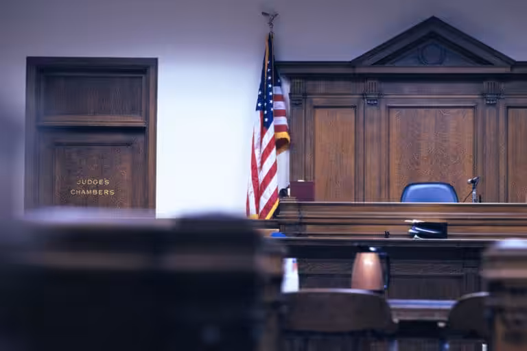 Empty courtroom with wooden judges bench, an American flag, and a door labeled Judges Chambers on the left. Several chairs and tables are visible in the foreground.