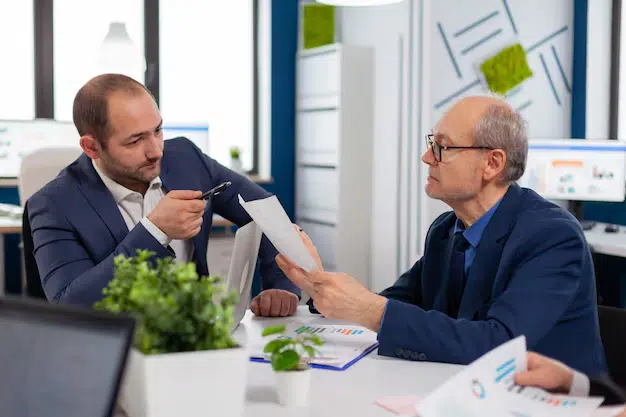 Two Men in Business Attire Sit at a Desk in an Office, Reviewing and Discussing Documents with Charts, Possibly Related to Midland Funding Llc. a Potted Plant and Office Supplies Are on the Table, While Others Work in the Background. - Ziegler Diamond Law sued by midland funding llc