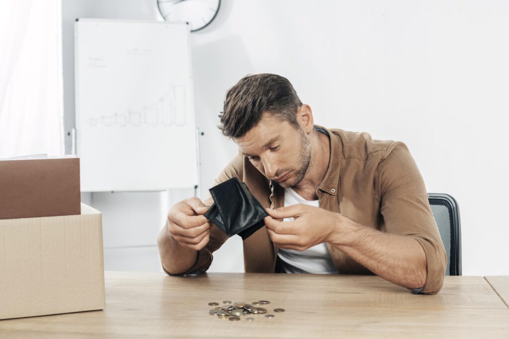 A man sits at a desk, looking into an empty wallet while coins are scattered on the table. He appears concerned about debt relief. In the background, there is a whiteboard with a graph and a clock on the wall.