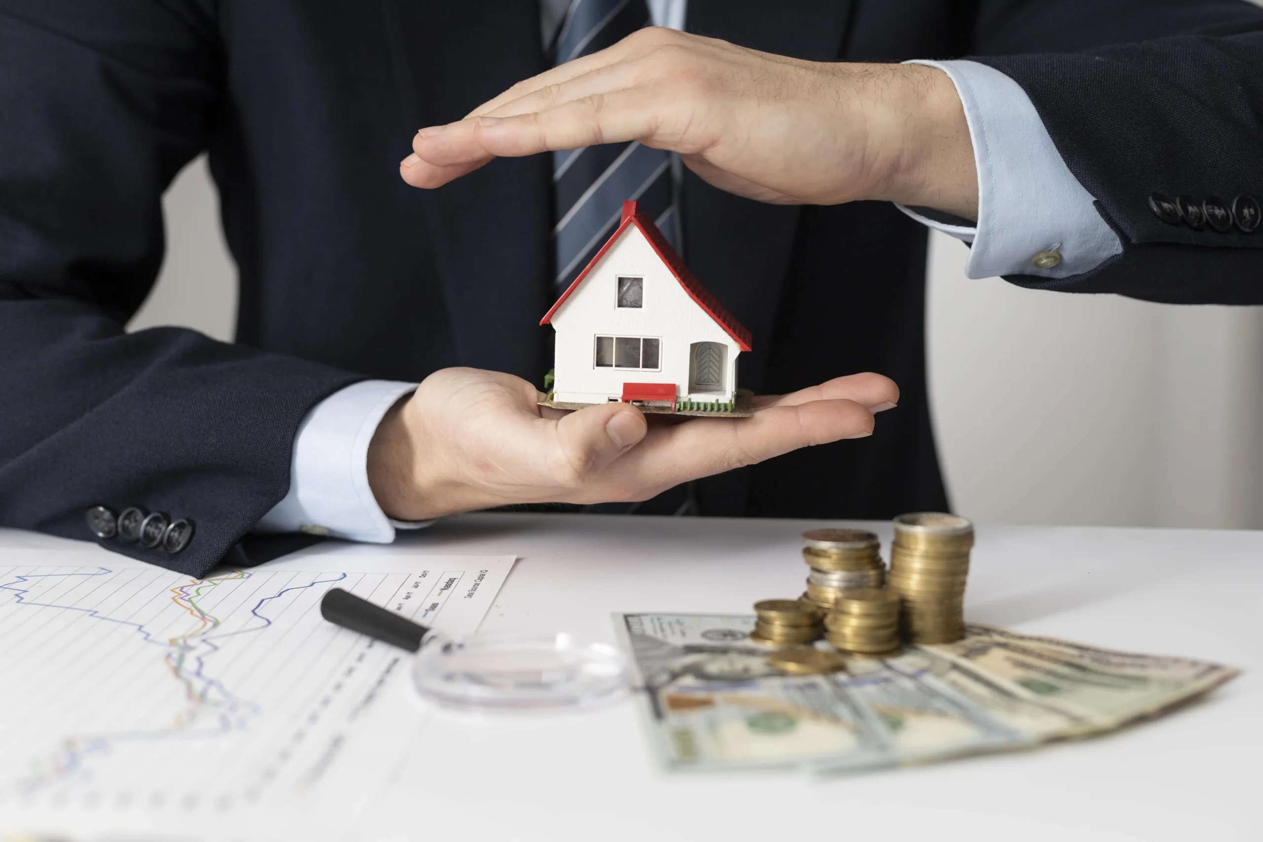 A person in a suit holds a small model house, with financial charts, coins, and bills on the table, hinting at buying a home after bankruptcy or exploring what is the best way to buy a house after bankruptcy.