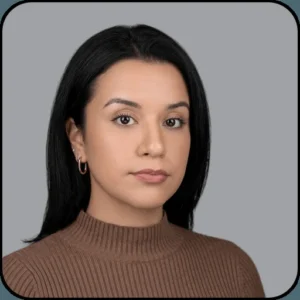A woman with straight black hair, wearing a brown ribbed turtleneck sweater and small hoop earrings, poses against a plain gray background. She has a neutral facial expression.