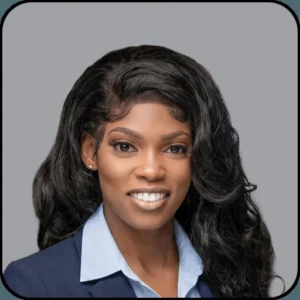 A woman with long, wavy black hair smiles at the camera. She is wearing a light blue collared shirt and a dark blazer, set against a plain gray background.