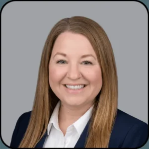 A woman with long, straight light brown hair smiles at the camera. She is wearing a white collared shirt and a dark blazer. The background is plain and light gray.