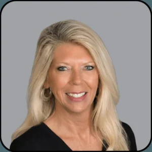 A woman with long, straight blonde hair and blue eyes is smiling at the camera. She is wearing hoop earrings and a black top, against a plain gray background.
