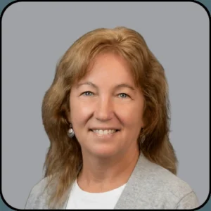 A woman with light brown, wavy hair and blue eyes is smiling. She is wearing a white top, a light-colored jacket, and dangle earrings. The background is a plain light gray.