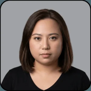 A person with straight, shoulder-length brown hair and a neutral expression, wearing a black shirt, poses against a plain gray background.