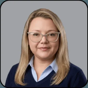 A woman with long blonde hair, wearing glasses, a light blue collared shirt, and a navy sweater, smiles softly against a plain gray background.