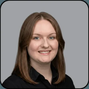 A woman with straight, shoulder-length brown hair and light skin smiles at the camera. She is wearing a black collared shirt and is photographed against a plain gray background.