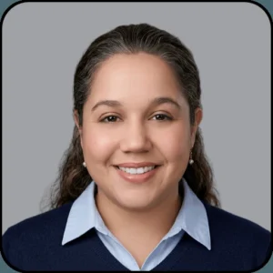 A woman with long dark hair, smiling, wearing a light blue collared shirt under a dark blue sweater, and small earrings, against a plain gray background.