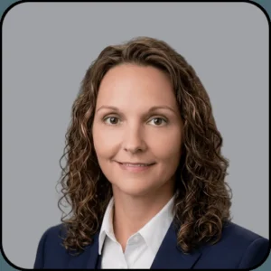 Portrait of a woman with long curly brown hair, wearing a navy blazer and white shirt, smiling slightly at the camera against a plain gray background.