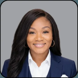 A woman with long dark hair, wearing a navy blue blazer and white shirt, smiles at the camera against a plain gray background.