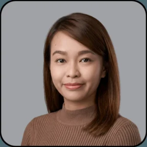 Portrait of a woman with straight, shoulder-length brown hair, wearing a ribbed brown sweater, smiling softly against a plain gray background.