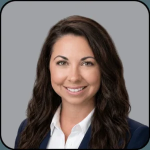 Professional headshot of a woman with long wavy brown hair, wearing a white collared shirt and dark blazer, smiling at the camera against a plain gray background.