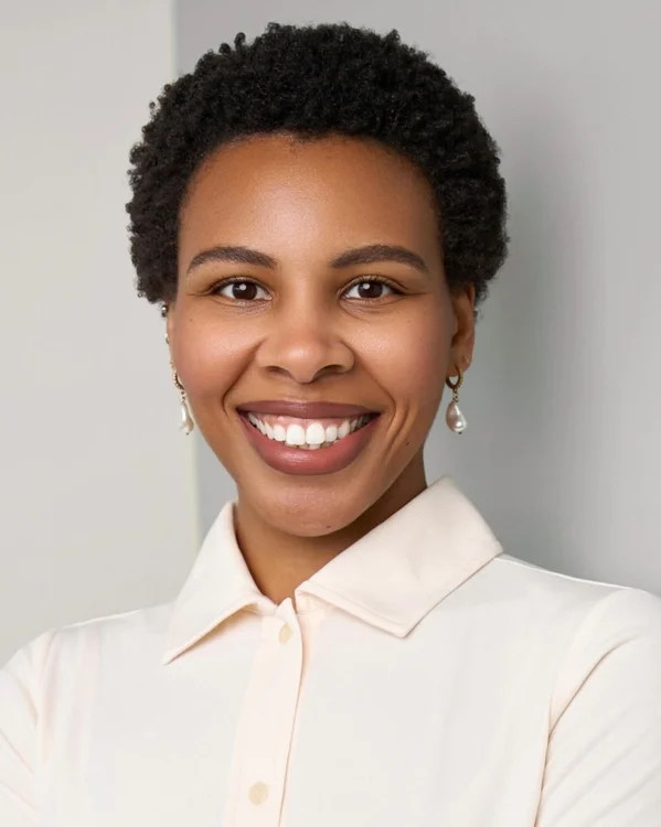 A woman with short curly hair smiles at the camera. She is wearing a cream-colored collared shirt and pearl earrings, standing against a light, neutral background.